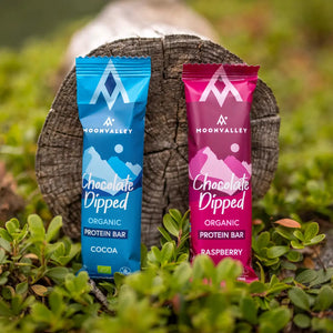 Moonvalley organic protein bars in Cocoa and Raspberry flavours, photographed in a natural outdoor setting.