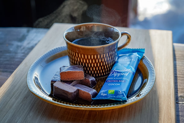 A cup of coffee and a Moonvalley chocolate protein bar on a wooden tray.