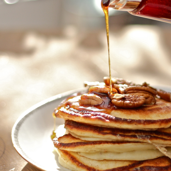 Stack of protein-rich pancakes with syrup and pecans being poured over them on a white plate.